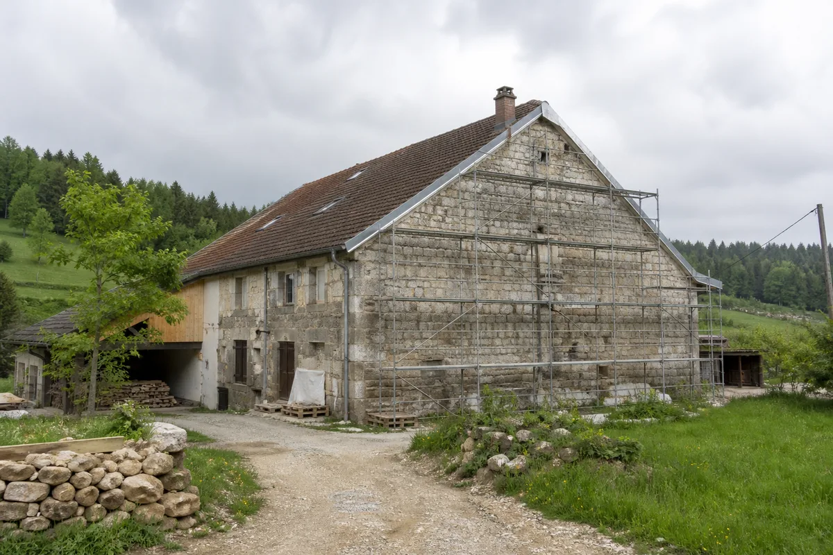 Corps de ferme jurassien en cours de rénovation, échafaudage posé le long du mur pignon en pierre